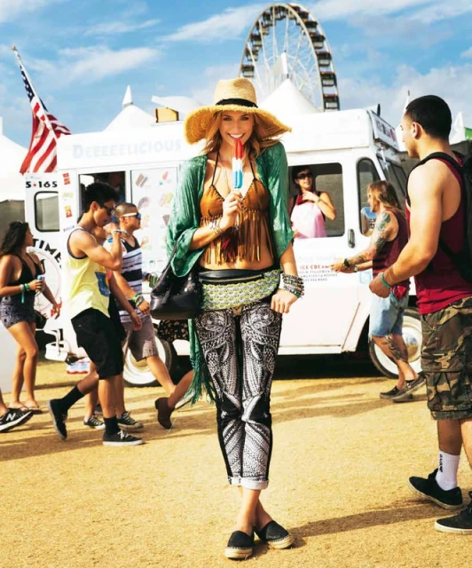 Outdoor shot of a woman in patterned leggings, a straw hat, and a bikini top, holding a popsicle, with food trucks in the background.