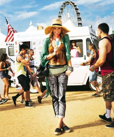 Outdoor shot of a woman in patterned leggings, a straw hat, and a bikini top, holding a popsicle, with food trucks in the background.