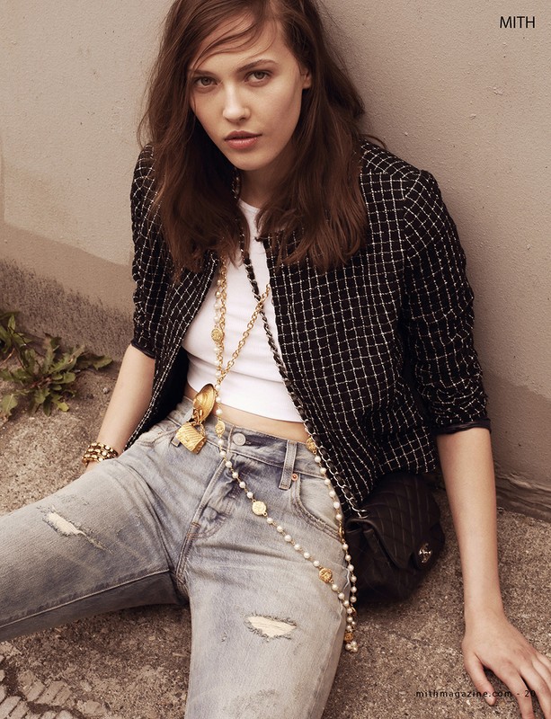 Fashion shot of a young adult woman wearing a white top, jeans, and a patterned jacket, accessorized with layered necklaces and a handbag.