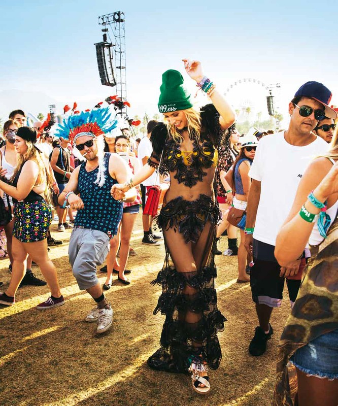 People celebrate and dance at an outdoor music festival in the daytime, with a woman in a feathered dress as the focal point.
