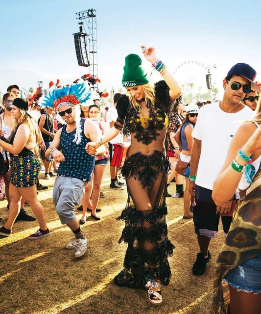 People celebrate and dance at an outdoor music festival in the daytime, with a woman in a feathered dress as the focal point.