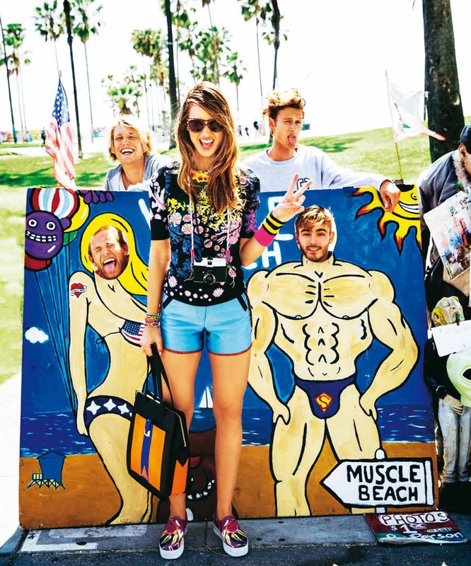 A young woman stands in front of a boardwalk art display, wearing colorful clothes, sunglasses, and an over-the-shoulder camera on a sunny day.