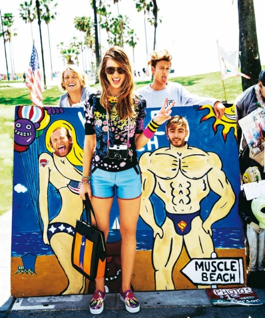 A young woman stands in front of a boardwalk art display, wearing colorful clothes, sunglasses, and an over-the-shoulder camera on a sunny day.