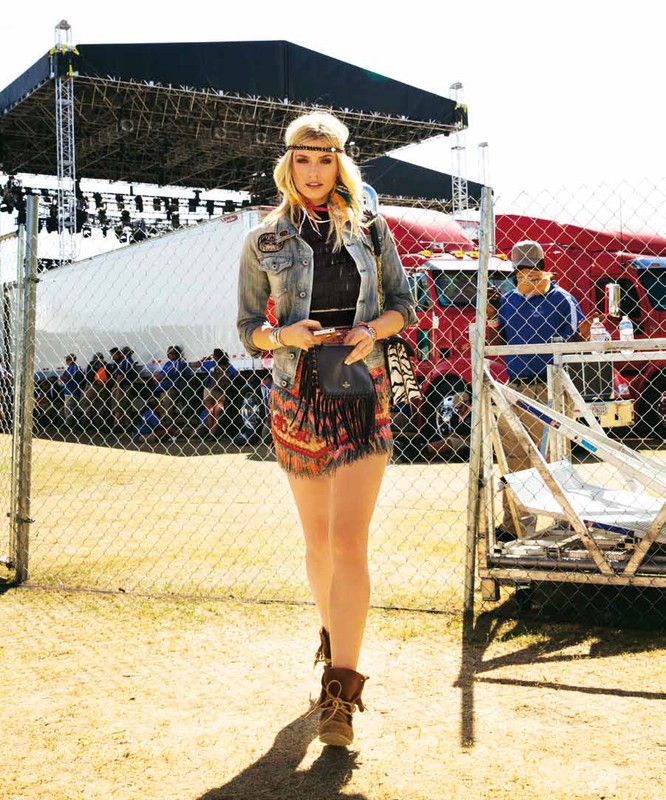 Young blonde woman in boho-style outfit walking towards camera at an outdoor festival, with stage and trucks visible in background.