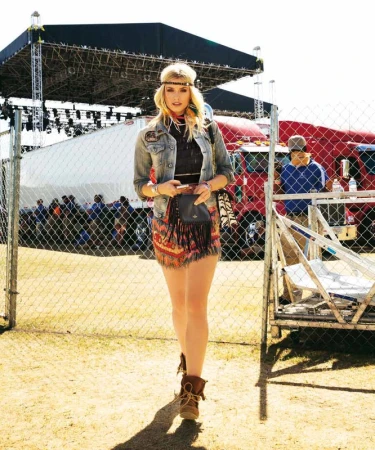 Young blonde woman in boho-style outfit walking towards camera at an outdoor festival, with stage and trucks visible in background.