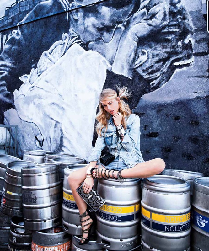Woman sits cross-legged on metal beer kegs in front of a mural wall. She wears denim jacket and shorts, with gladiator-style heeled sandals.