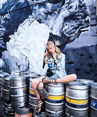 Woman sits cross-legged on metal beer kegs in front of a mural wall. She wears denim jacket and shorts, with gladiator-style heeled sandals.