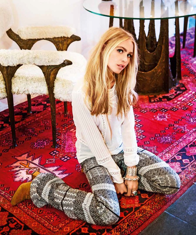 A fair-skinned woman with long blonde hair sits on a red patterned rug wearing a white top and patterned pants in an indoor setting.