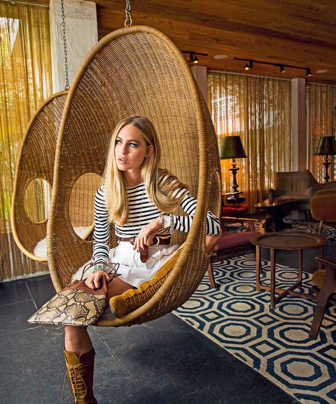 Stylish blonde woman in a rattan hanging chair, wearing a striped shirt and white skirt, accessorized with brown belt and boots indoors.