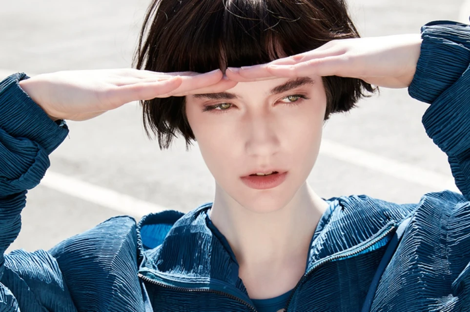 Eye-level close-up of a young adult female model in a blue crinkled jacket holding her hands to her brow in an outdoor setting.