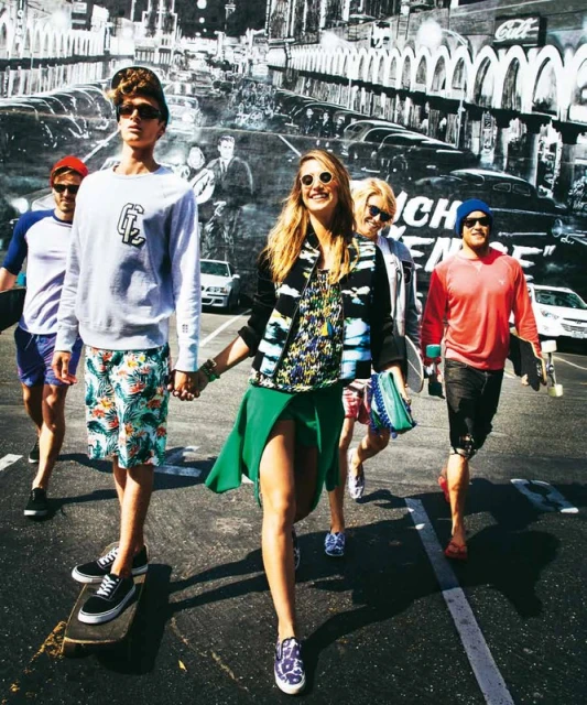 Group of young adults walks across a black asphalt street with white lines in sunny weather.  Street art in the background.