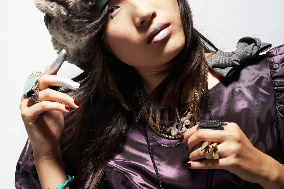 Close-up of a stylish woman wearing a fur hat, purple dress, and jewelry against a white background in a beauty and fashion shot.