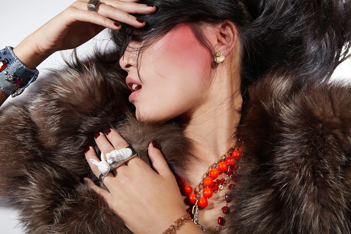 Fashion portrait of a woman wearing a fur coat, statement jewelry, and vibrant makeup in a studio setting against a white backdrop.