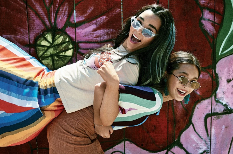 Two happy adult women pose back-to-back outdoors in front of a mural featuring a painted flower. The women wear sunglasses and colorful outfits.