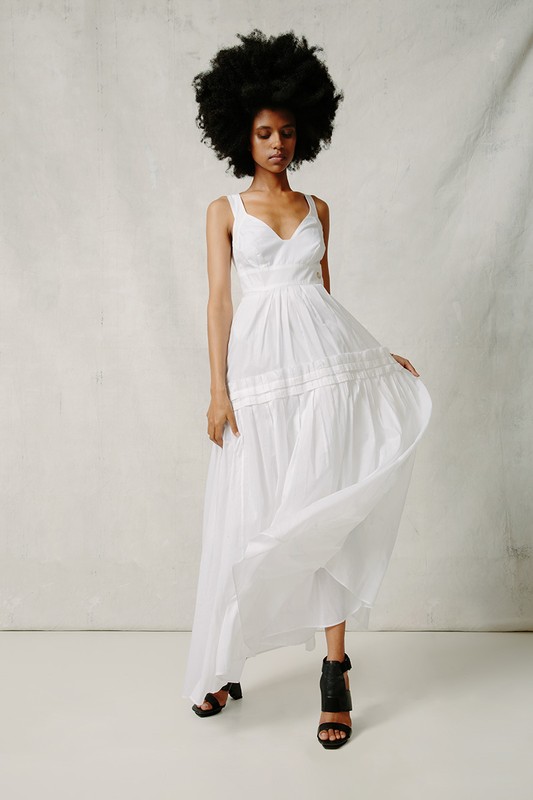 A woman with an afro poses in a flowing white dress, accessorized with block heel sandals. The scene is set against a simple backdrop.