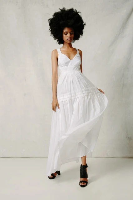 A woman with an afro poses in a flowing white dress, accessorized with block heel sandals. The scene is set against a simple backdrop.