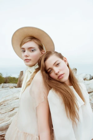 Two young women with long brown hair leaning back to back outdoors with light clothing and a wide-brimmed hat