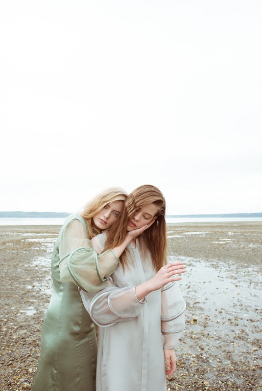 Two female models in long dresses standing on a pebble shore by water with hair styling by Erin Skipley