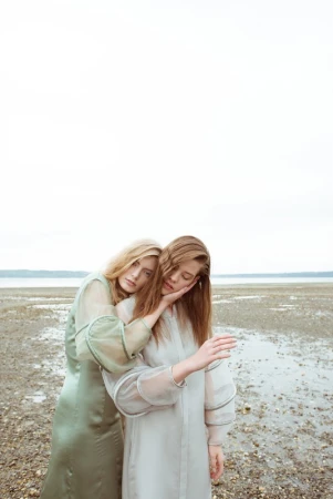 Two female models in long dresses standing on a pebble shore by water with hair styling by Erin Skipley