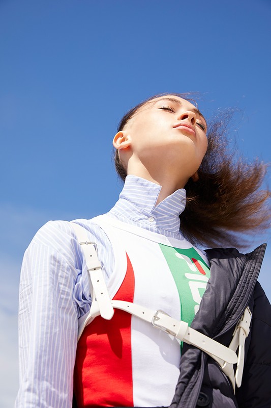 Woman in high-fashion sportswear, shot from low angle, contrasts a white top with red and green accents against a clear blue sky.