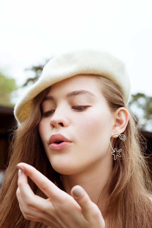 Portrait of a young woman with light skin, wearing a cream-colored beret and a star earring, blowing a kiss toward the camera.
