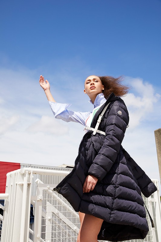 A woman poses in a stylish blue puffer coat against a sunny blue sky. She is wearing a collared shirt and white harness.