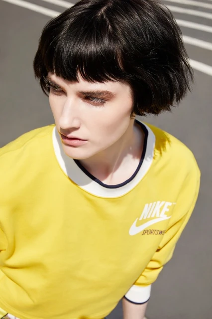 Woman with short dark hair wears a yellow Nike Sportswear sweatshirt in an outdoor shot with white shadows. Fashion and portrait photography.