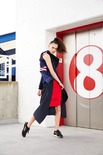 Fashionable adult female in a red dress, windbreaker, and sneakers, standing near a metal door and concrete wall in a daytime shot.