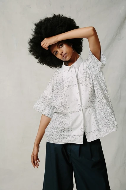 A black woman poses indoors wearing a white eyelet shirt and wide-legged pants. She has dark curly hair and is looking at the camera.