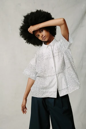 A black woman poses indoors wearing a white eyelet shirt and wide-legged pants. She has dark curly hair and is looking at the camera.