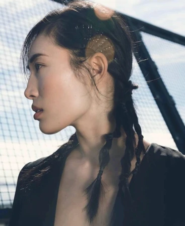 Profile portrait of a woman with braided hair and bobby pin details against an outdoor mesh backdrop