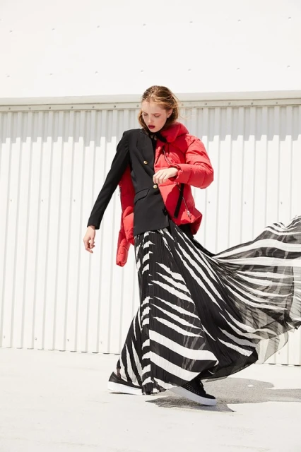 Fashion shot of a young woman in a red puffer jacket and black and white skirt, posing against a white wall in an outdoor setting.
