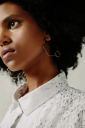 Close-up portrait of a woman with dark curly hair wearing sculptural gold hoop earrings and a speckled white shirt