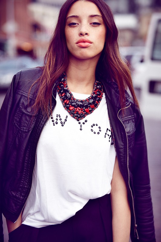 A woman in a leather jacket and t-shirt with a statement necklace poses against a blurred urban backdrop with parked cars and buildings.