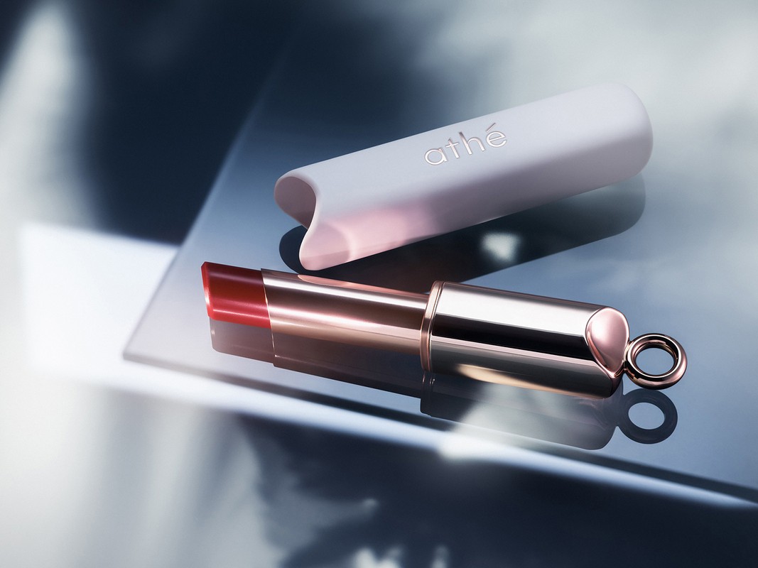 Athe lipstick tube with red balm and rose gold metallic base resting on a reflective mirror surface next to its white cap