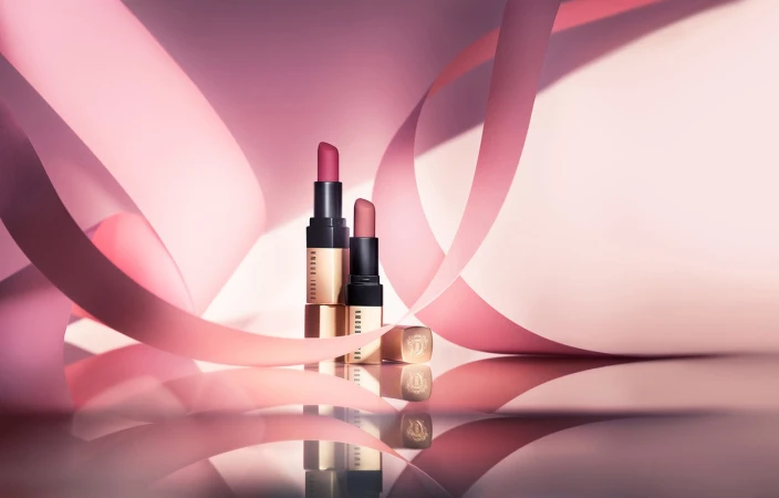 Two lipstick tubes standing upright surrounded by curved pink ribbons on a reflective surface