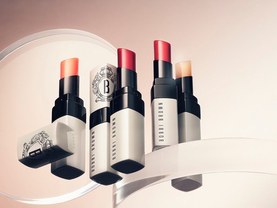 Close-up of several Bobbi Brown lipsticks in various shades on a clear stand against a soft gradient peach background.