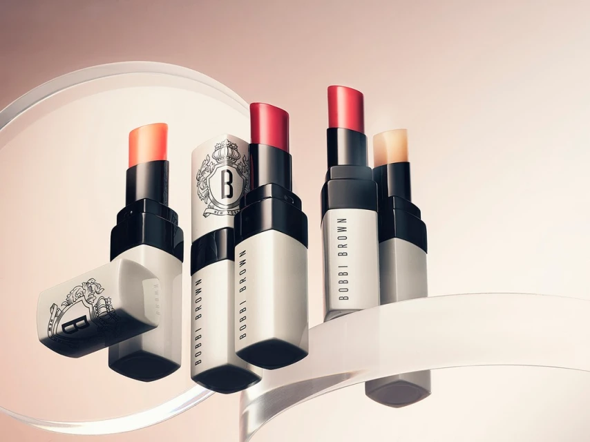 Close-up of several Bobbi Brown lipsticks in various shades on a clear stand against a soft gradient peach background.