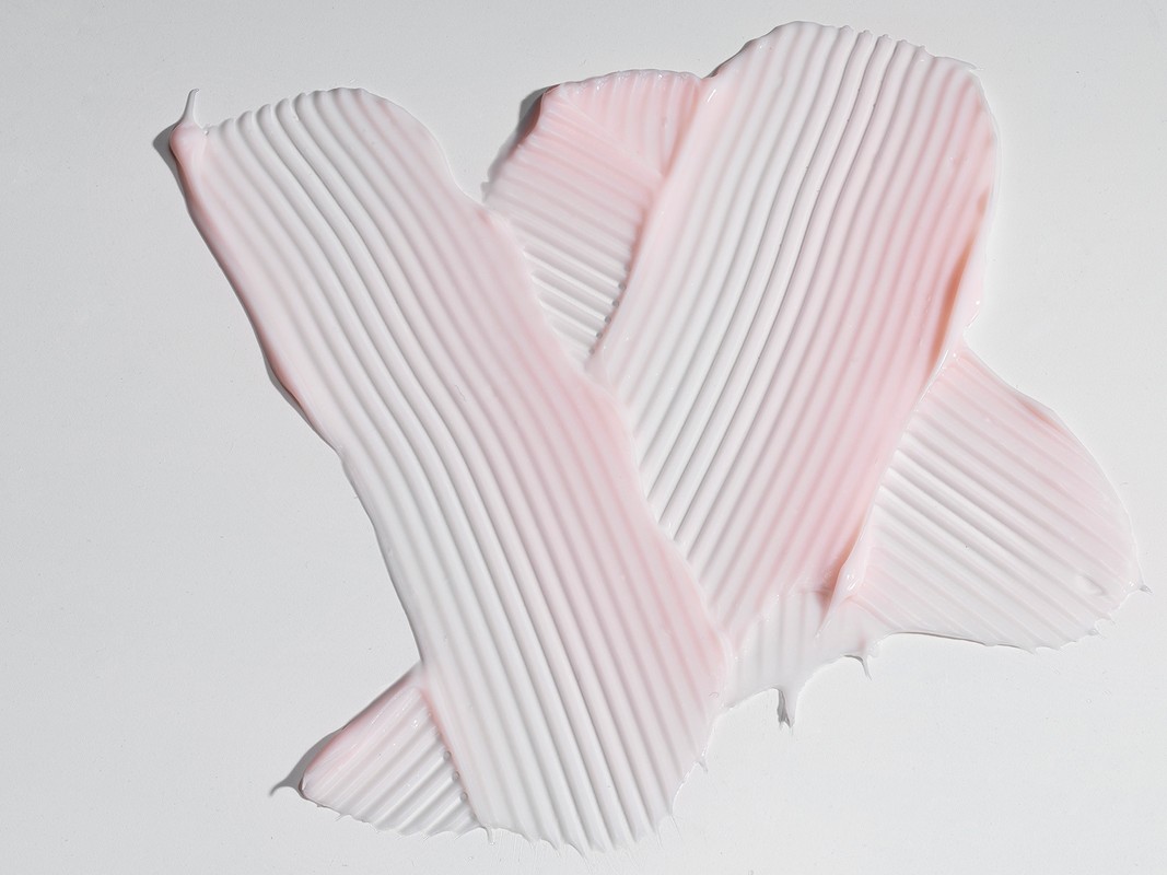 Close-up of a thick cream with parallel ridges swiped across a white background, creating a textured pattern with pink and white hues.