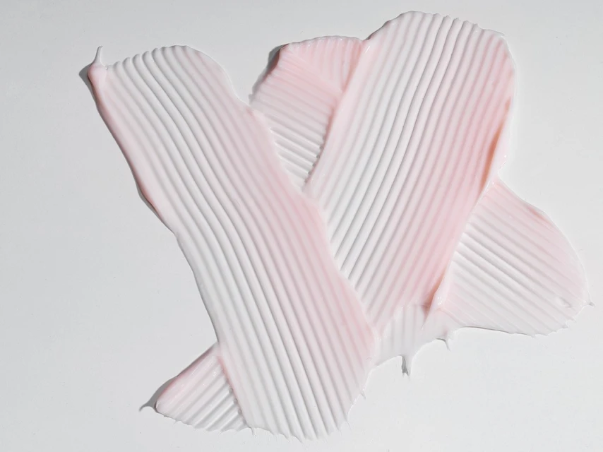 Close-up of a thick cream with parallel ridges swiped across a white background, creating a textured pattern with pink and white hues.