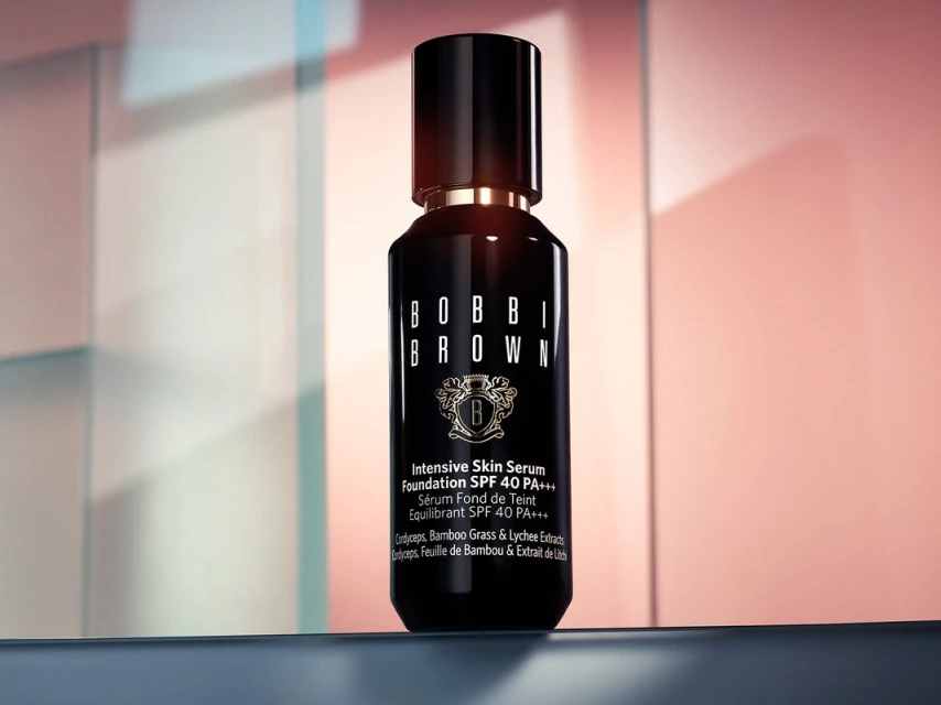 Bobbi Brown Intensive Skin Serum Foundation in a sleek black bottle against a blurred background with coral and blue lighting.