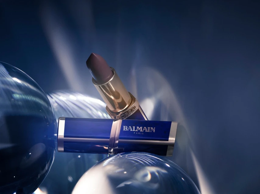 L'Oreal Paris and Balmain collaboration lipstick tube in a gold case with a mauve lipstick bullet resting on a blue tube and clear sphere.