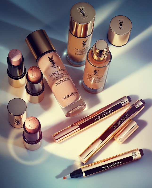 Still life of Yves Saint Laurent makeup products in neutral and gold tones, featuring foundation, highlighter sticks, and concealer pens.