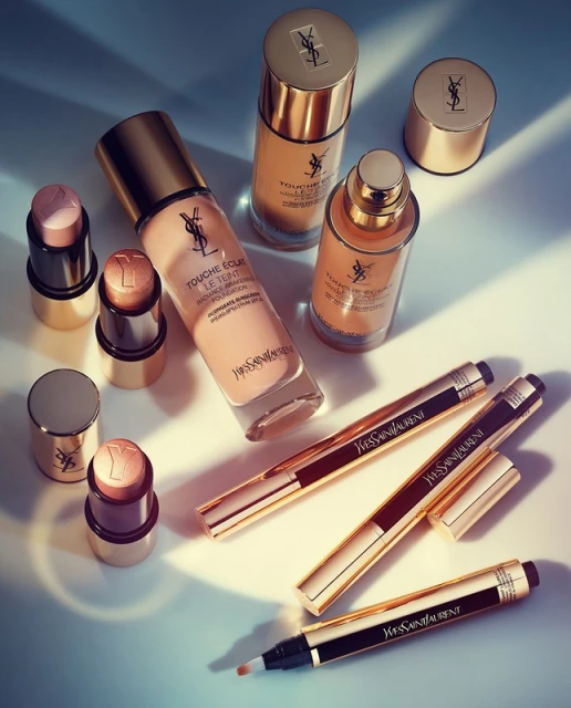 Still life of Yves Saint Laurent makeup products in neutral and gold tones, featuring foundation, highlighter sticks, and concealer pens.