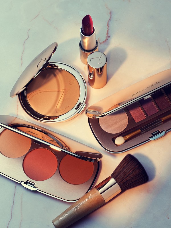 Close-up of opened and closed makeup compacts, a lipstick tube, and a makeup brush arranged on a white marble surface.