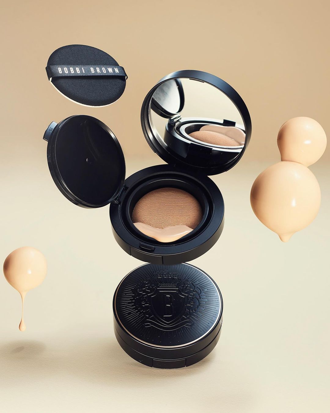 Cosmetic product arrangement featuring a black compact case, makeup sponge, and droplets of liquid foundation on a beige background.