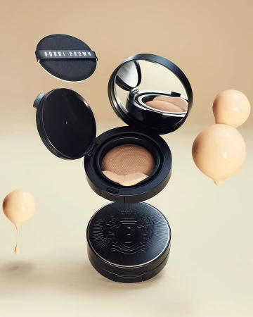 Cosmetic product arrangement featuring a black compact case, makeup sponge, and droplets of liquid foundation on a beige background.