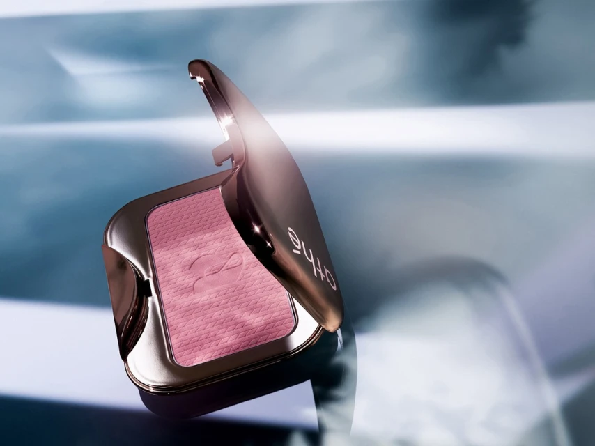Cosmetic powder compact with reflective rose-gold case and pink powder is shown in a beauty shot against a blurred blue background.