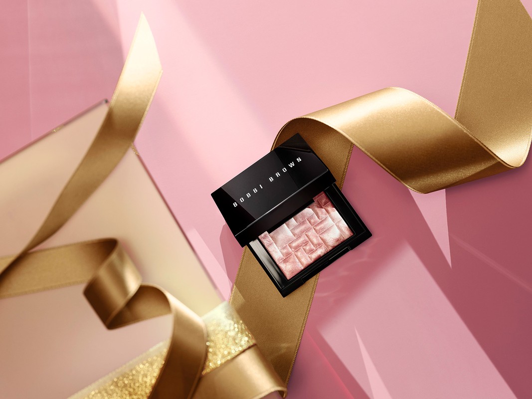 Close-up of Bobbi Brown makeup compact on a pink surface, accented by flowing gold satin ribbon. Beauty and cosmetic product shot.