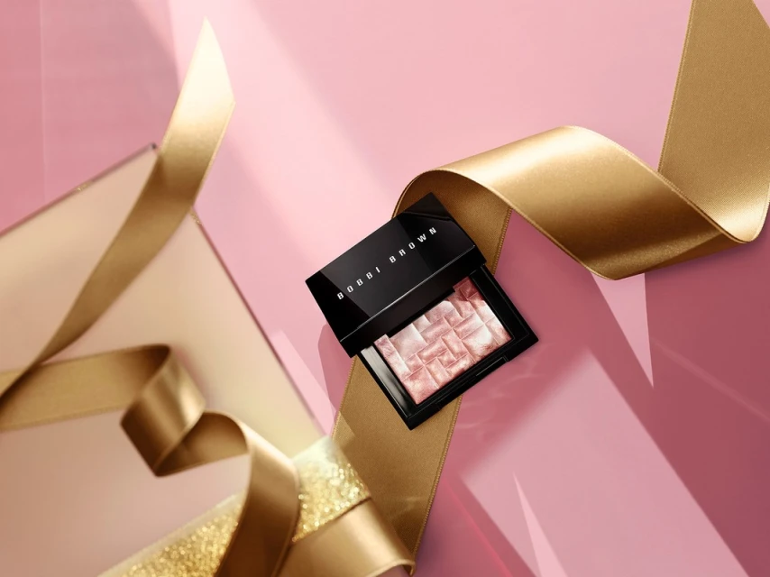 Close-up of Bobbi Brown makeup compact on a pink surface, accented by flowing gold satin ribbon. Beauty and cosmetic product shot.